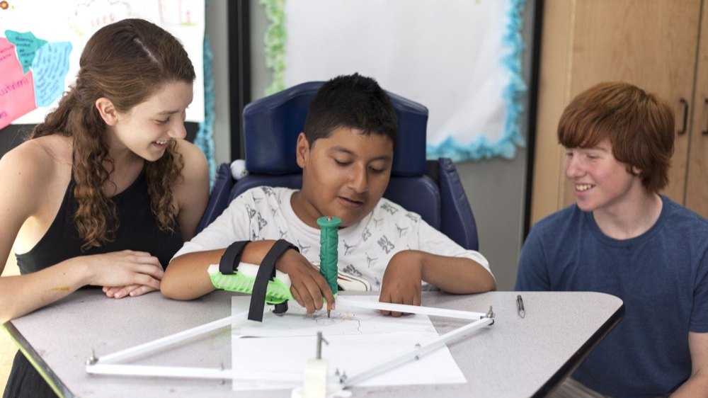 Two students at a table with a child who uses a wheelchair, fitting a device they made for him